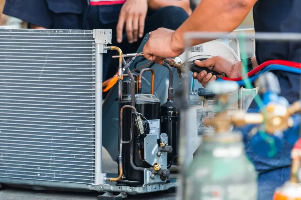 Hands working on the copper lines and compressor of an opened outdoor air conditioning unit, with gas cylinders in the foreground.