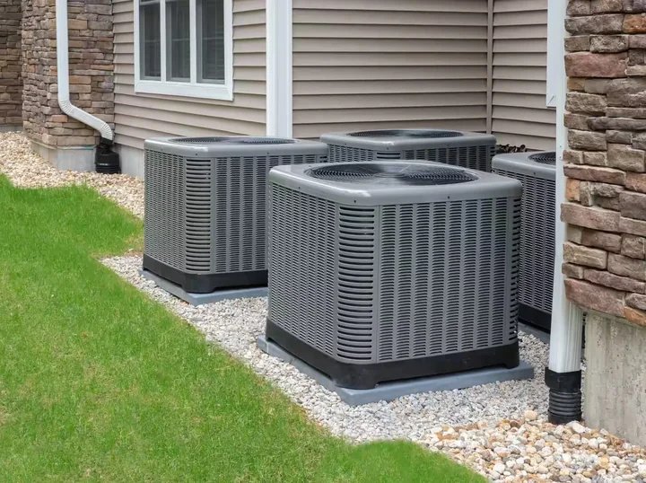 Three outdoor air conditioner condenser units on a gravel bed beside a house with tan siding and stone trim.